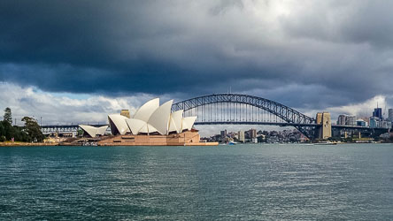 A wide landscape view across Sydney Harbour on a stormy day, featuring the Sydney Opera House with its white sail roofs in the foreground and the steel arch of the Sydney Harbour Bridge in the background under heavy grey clouds.